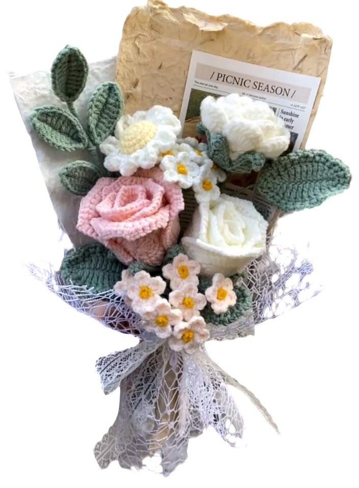 A handmade crochet bouquet with a pink rose, white daisies, and green leaves wrapped in white lace