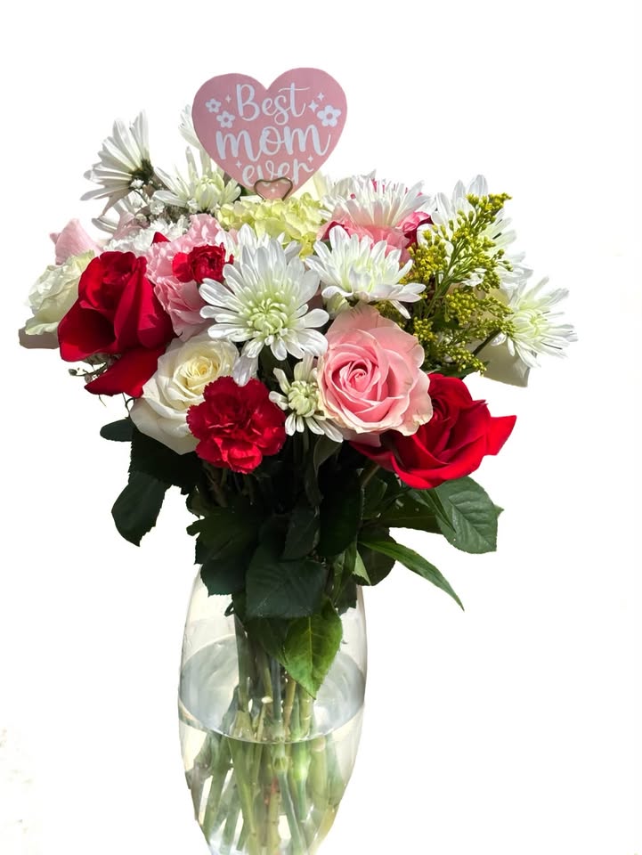 A mixed arrangement of red roses, pink roses, white chrysanthemums and carnations in a glass vase with a Best Mom Ever heart pick